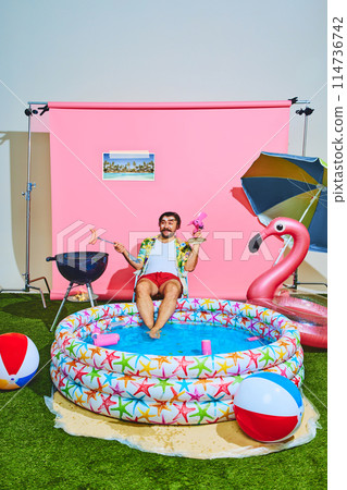 Young man in vibrant outfit with tropical shirt relaxing in inflatable swimming pool and cooking barbeque-sausages. Young man in vibrant outfit with tropical shirt relaxing in inflatable swimming pool and cooking barbeque-sausages. 114736742