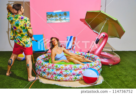 Rear view of man in tropical shirt and red shorts have fun spraying water at woman relaxing in inflatable pool Rear view of man in tropical shirt and red shorts have fun spraying water at woman relaxing in inflatable pool 114736752
