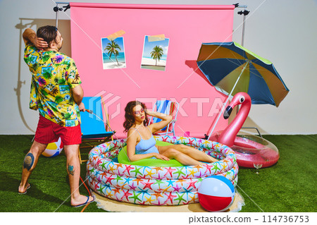Young man spraying water from hose at beautiful woman in vibrant kiddie pool. Fun and festive summer vibe. Young man spraying water from hose at beautiful woman in vibrant kiddie pool. Fun and festive summer vibe. 114736753