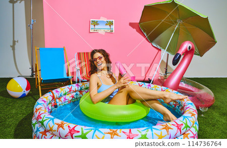 Lounging in colorful inflatable pool, slim young woman enjoys sun on neon-green float ring and drinks cold, refreshing soda. Lounging in colorful inflatable pool, slim young woman enjoys sun on neon-green float ring and drinks cold, refreshing soda. 114736764