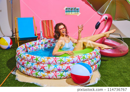 Young beautiful woman enjoying her staycation in kiddie pool swimming in inflatable ring and drinking cocktail. 114736767