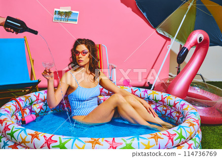 Woman relaxes in colorful kiddie pool with cocktail in hand, while waiter pours drink. Beach retreat. Contemporary colors. Woman relaxes in colorful kiddie pool with cocktail in hand, while waiter pours drink. Beach retreat. Contemporary colors. 114736769