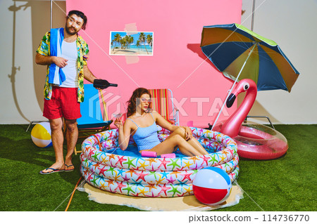 Man, wearing tropical shirt and red shorts, pours drink while woman, lounging in inflatable pool in striped swimsuit, holds cocktail glass. 114736770