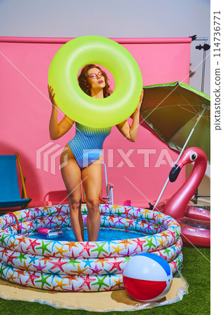 Woman enjoying playful moment in inflatable pool, holding neon-green swim ring against pink studio background. Woman enjoying playful moment in inflatable pool, holding neon-green swim ring against pink studio background. 114736771