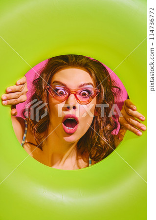 Portrait of surprised young woman holds neon-green inflatable ring, peeking out of it against bright pink background. 114736772