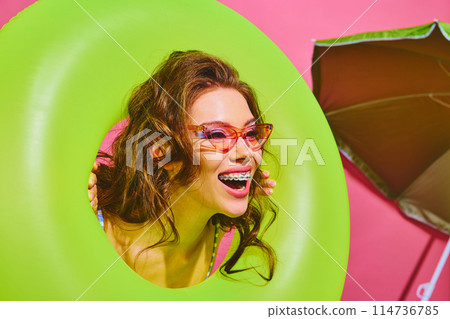 Portrait of young woman holds neon-green inflatable ring and smiles brightly peeking out of it against bright pink background. Portrait of young woman holds neon-green inflatable ring and smiles brightly peeking out of it against bright pink background. 114736785