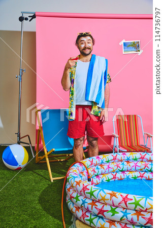 Man dressed vibrant shorts and shirt holding refreshing cocktail standing near inflatable pool against studio background. 114736797