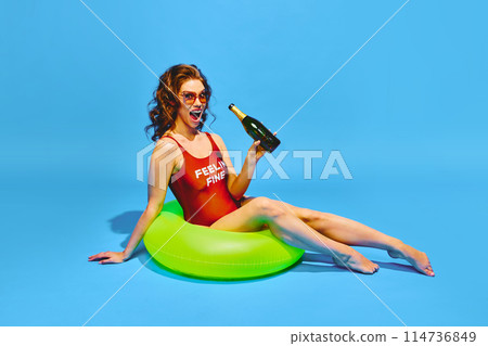 Happy young woman dressed in retro swimsuit drinks champagne resting in neon colored inflatable ring against blue background. Happy young woman dressed in retro swimsuit drinks champagne resting in neon colored inflatable ring against blue background. 114736849