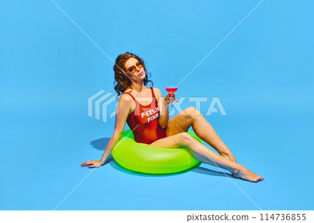 Beautiful, slim woman in retro swimwear sitting in neon colored inflatable ring with cocktail against blue background. Beautiful, slim woman in retro swimwear sitting in neon colored inflatable ring with cocktail against blue background. 114736855