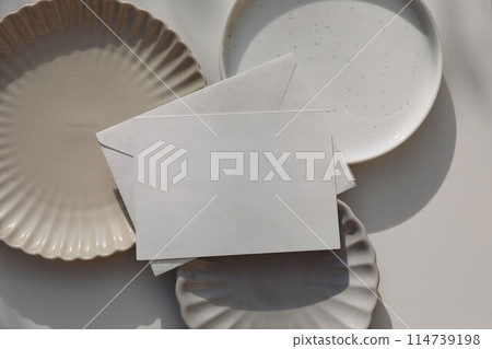 Empty horizontal greeting card, invitation mockup, envelope in sunlight. Various plates on beige table background. Soft shadows. Neutral still life, web banner. Food, restaurant, craft concept, flat 114739198