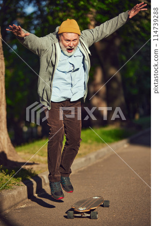 Elderly man in casual stylish clothes skateboarding in morning park on sunny day, training stunts. Youthful activity Elderly man in casual stylish clothes skateboarding in morning park on sunny day, training stunts. Youthful activity 114739288