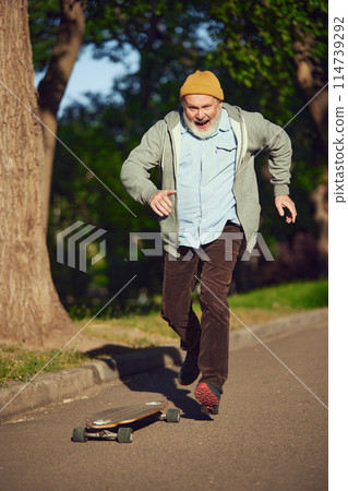 Elderly man in casual stylish clothes skateboarding in morning park on sunny day, training stunts. Youthful activity Elderly man in casual stylish clothes skateboarding in morning park on sunny day, training stunts. Youthful activity 114739292