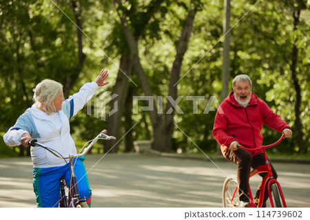 Cycling Activity. Elderly couple, man and woman, riding bicycles in lush park on warm summer day. Emotional happy people Cycling Activity. Elderly couple, man and woman, riding bicycles in lush park on warm summer day. Emotional happy people 114739602