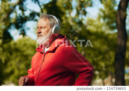 Senior man jogging in park, in red hoodie and headphones. Early morning running routine. Outdoor training Senior man jogging in park, in red hoodie and headphones. Early morning running routine. Outdoor training 114739603