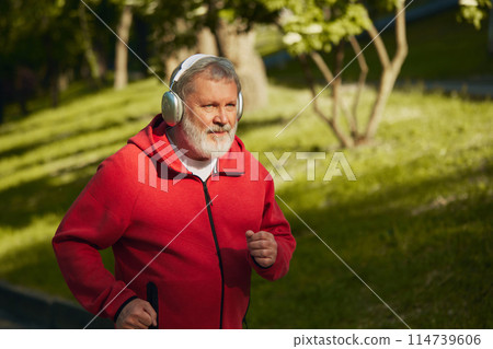 Morning Run. Elderly man in red sportswear and headphones jogging in park on early sunny morning. Outdoor activity. Morning Run. Elderly man in red sportswear and headphones jogging in park on early sunny morning. Outdoor activity. 114739606