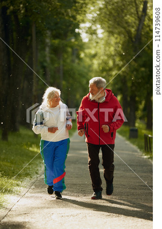 Elderly and active man and woman, positive couple in sportswear energetically jogging in early morning park, training. 114739608