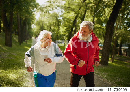 Senior Fitness Enthusiasts. Lively elderly couple, man and woman in sportswear running in park in the morning, growing energy and keeping toned Senior Fitness Enthusiasts. Lively elderly couple, man and woman in sportswear running in park in the morning, growing energy and keeping toned 114739609