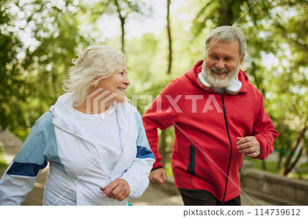 Sport inspirational. Happy smiling elderly people, man and woman in sportswear jogging in public park in early morning. 114739612