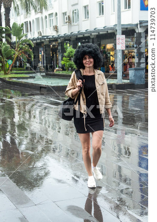 A young woman of 30 years old walks in the rain along a small town street 114739703