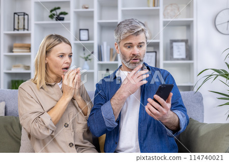 Middle-aged couple with shocked expressions, looking at a smartphone while sitting on the couch in a modern living room. 114740021