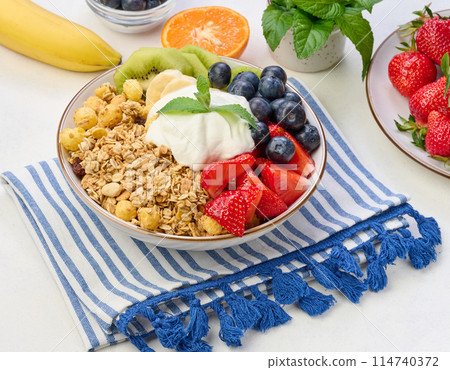 Granola with strawberries, kiwi, banana and blueberries in a round plate on a white table. Granola with strawberries, kiwi, banana and blueberries in a round plate on a white table. 114740372