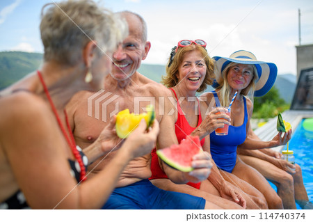 Group of cheerful seniors sitting on edge of pool with legs in water, eating watermelon. Elderly friends spending hot day by swimming pool, sunbathing. Group of cheerful seniors sitting on edge of pool with legs in water, eating watermelon. Elderly friends spending hot day by swimming pool, sunbathing. 114740374