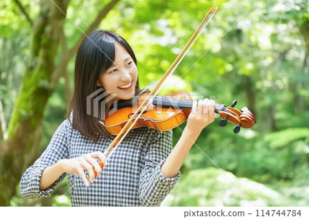 A woman playing the violin with a forest of fresh green leaves in the background A woman playing the violin with a forest of fresh green leaves in the background 114744784