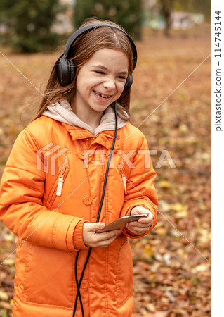 Laughing cheerful girl listens to music on headphones in an autumn park. 114745144