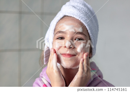 A girl in the bathroom with a white towel on her head washes her face with cleansing foam, smiles 114745161