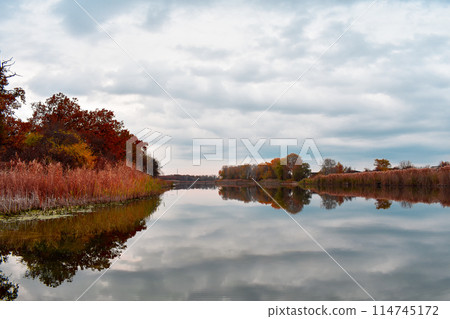 Lake in autumn, surrounded by trees on the shore in orange and red leaves under a cloudy sky 114745172