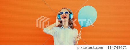 happy cheerful young woman in headphones listening to music with blue balloon on orange background 114745671