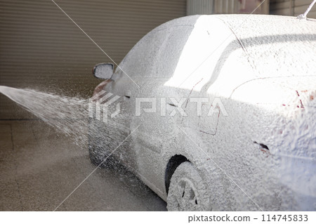 White foam from a sprayer flies onto a car at a car wash. The driver covers the entire car with soap foam to wash away the dirt. Car wash at a self-service service. White foam from a sprayer flies onto a car at a car wash. The driver covers the entire car with soap foam to wash away the dirt. Car wash at a self-service service. 114745833