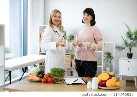 Mature woman in doctor's coat finding out waist length of female patient. 114746025