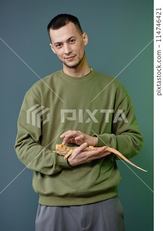 Vertical portrait of Asian young man holding iguana in hands carefully standing against green background and looking at camera Vertical portrait of Asian young man holding iguana in hands carefully standing against green background and looking at camera 114746421