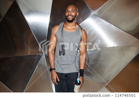 Waist up portrait of muscular Black man smiling at camera standing against graphic metal wall in gym, shot with flash 114746501