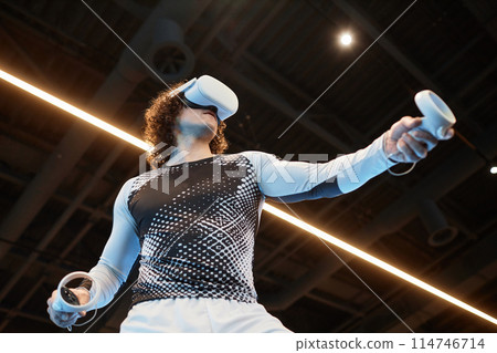 Futuristic low angle shot of man wearing VR headset in gym and training against neon light strip copy space Futuristic low angle shot of man wearing VR headset in gym and training against neon light strip copy space 114746714