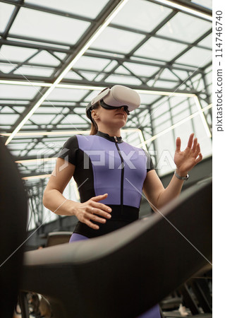 Vertical low angle portrait of sportive woman running on treadmill and wearing VR headset in futuristic gym 114746740