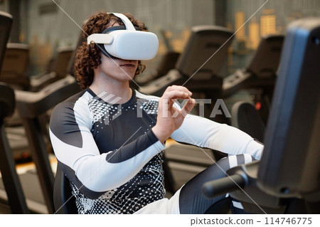 Portrait of young man working out with exercise bike in gym and wearing VR headset gesturing and enjoying immersive training experience Portrait of young man working out with exercise bike in gym and wearing VR headset gesturing and enjoying immersive training experience 114746775