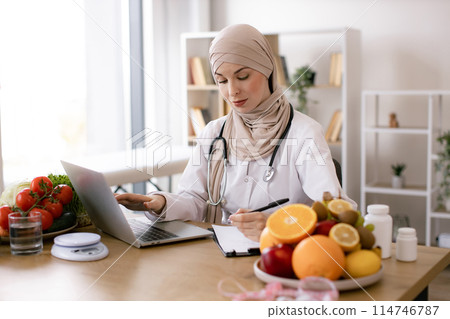Muslim female nutritionist sitting and working with laptop in modern clinic. 114746787