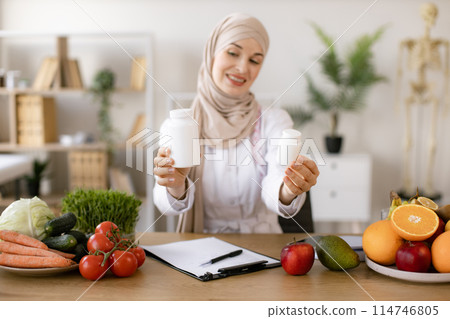 Portrait of positive Muslim lady in hijab and lab coat smiling at camera. Portrait of positive Muslim lady in hijab and lab coat smiling at camera. 114746805