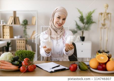 Portrait of positive Muslim lady in hijab and lab coat smiling at camera. 114746807