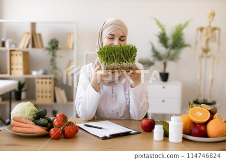 Lady in white coat holding container with microgreens while sitting at desk. Lady in white coat holding container with microgreens while sitting at desk. 114746824