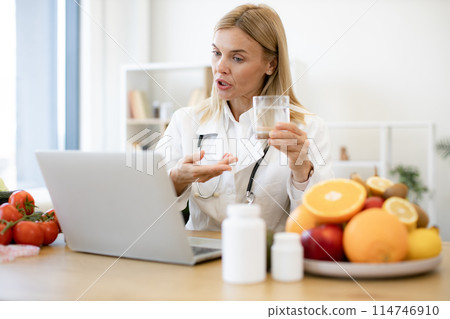 Female doctor in white coat holding glass of water during a video call. Female doctor in white coat holding glass of water during a video call. 114746910