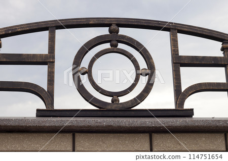 Close-up detail of beautiful vintage forged metal big dark abstract decoration on top of stone granite gray brown fence. Work of blacksmith, protection, art and fantasy concept. Close-up detail of beautiful vintage forged metal big dark abstract decoration on top of stone granite gray brown fence. Work of blacksmith, protection, art and fantasy concept. 114751654