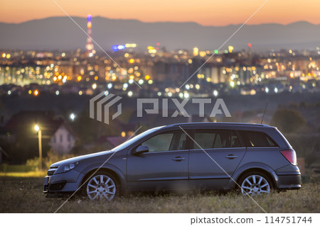 Gray car parked at night in green meadows on background of lights of distant city buildings and dark mountain ridge under bright sky at sunset Gray car parked at night in green meadows on background of lights of distant city buildings and dark mountain ridge under bright sky at sunset 114751744