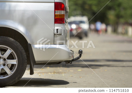 Close-up side view detail of silver passenger medium size luxury minibus van with tow-bar parked on summer sunny city street pavement with blurred silhouettes of pedestrians and cars in background. Close-up side view detail of silver passenger medium size luxury minibus van with tow-bar parked on summer sunny city street pavement with blurred silhouettes of pedestrians and cars in background. 114751810