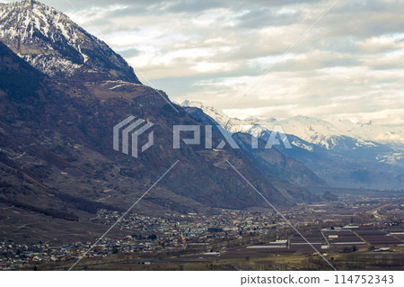 Small town in valley at the foot of magnificent Alps mountain, Switzerland. Quiet life on background of fantastic peaks covered with shiny white snow under bright blue sky with puffy white clouds. Small town in valley at the foot of magnificent Alps mountain, Switzerland. Quiet life on background of fantastic peaks covered with shiny white snow under bright blue sky with puffy white clouds. 114752343