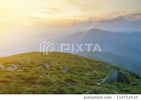 Summer camping in mountains at dawn. Tourist tent on round grassy hill on distant misty blue mountains range under pink sky before sunrise or sunset. Tourism, hiking and beauty of nature concept. Summer camping in mountains at dawn. Tourist tent on round grassy hill on distant misty blue mountains range under pink sky before sunrise or sunset. Tourism, hiking and beauty of nature concept. 114752410