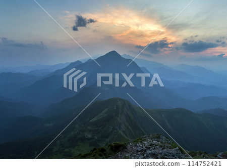 Fantastic sunrise or sunset over green mountain ridge covered with dense blue fog. Bright orange sun raising in soft cloudy sky over distant horizon. Beauty of nature, tourism and traveling concept. 114752421