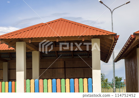 New modern alcove on kindergarten playground with bright orange tiling roof on blue sky background. New modern alcove on kindergarten playground with bright orange tiling roof on blue sky background. 114752452